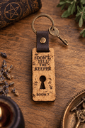Dont Tell The Keeper keychain in engraved pale wood with dark leather loop, brass-tone ring, keyhole cutout and Room 7 text
